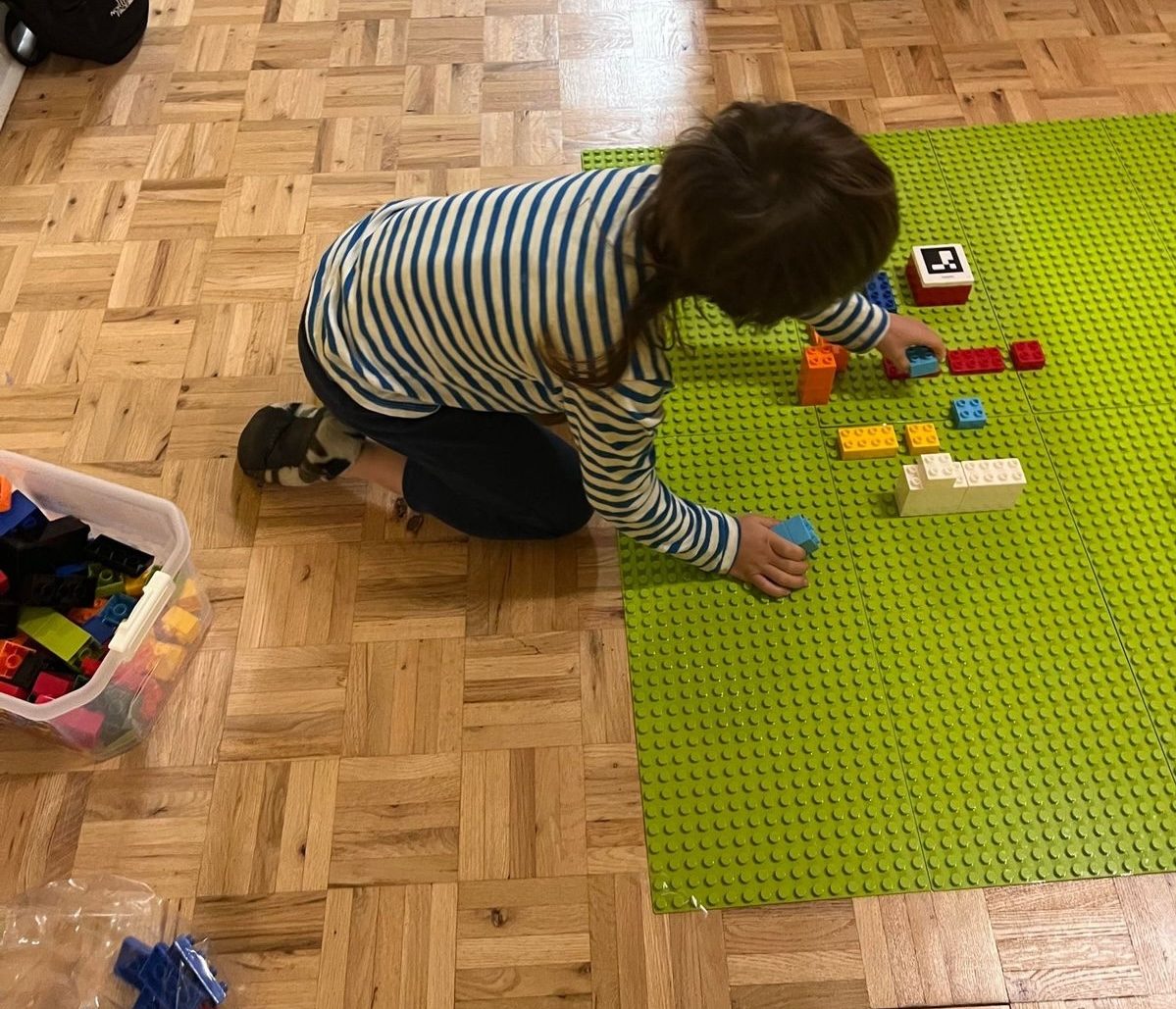 My 8-year-old, seen from behind, placing bricks on four green baseplates on the living-room floor