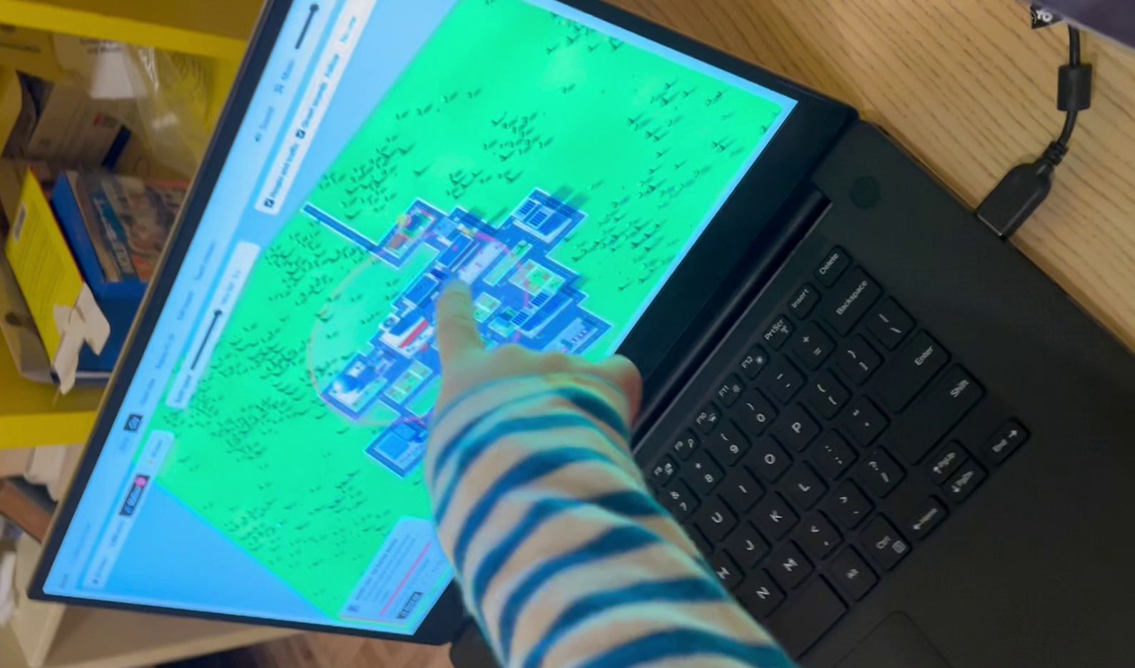 A child's arm in a striped shirt points at the 3D city on a laptop screen
